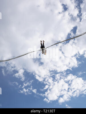 Girl hanging in slingshot against blue cloudy sky. Catapult exciting attraction in action. Girl enjoying her catapult amusement ride high-jumped to wh Stock Photo