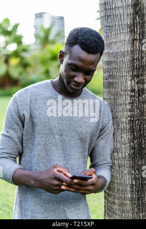 Young man leaning on tree trunk in a park, using smartphone Stock Photo