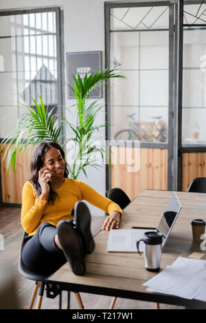 Young businesswoman sitting with feet on desk talking on cell phone Stock Photo