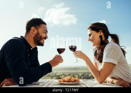 Side view of a couple sitting in an open air restaurant drinking red wine. Smiling couple toasting wine sitting at a restaurant. Stock Photo