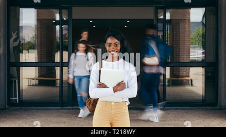 Portrait of girl student standing at university campus with other students walking in background. Young woman standing in college with students walkin