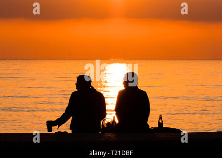 Aberystwyth, Wales. 29th Mar 2019. UK Weather: A couple of people enjoying an evening drinkon the promenade in Aberystwyth, on the Cardigan Bay coast of west Wales are silhouetted by the flaming setting sun . High pressure continues to dominate the weather for much England and Wales, with settled conditions forecast to last at least another day , before more cooler conditions return. Photo:Keith Morris/Alamy Live News Stock Photo