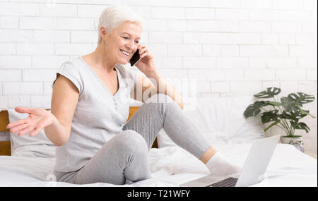 Home office. Successful senior woman sitting on bed working on laptop and talking on phone, panorama, free space Stock Photo