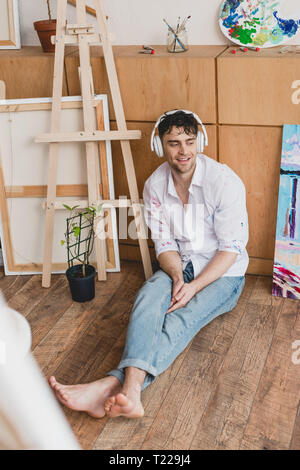 smiling artist in headphones, listening music while sitting on wooden floor Stock Photo