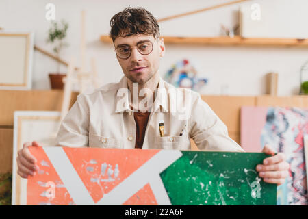 selective focus of handsome artist in eyeglasses holding picture and looking at camera Stock Photo
