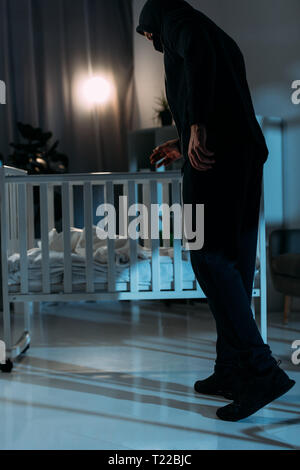 Kidnapper in black clothes standing near crib in dark room Stock Photo