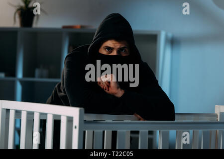 Worried kidnapper in black clothes and mask standing beside crib Stock Photo