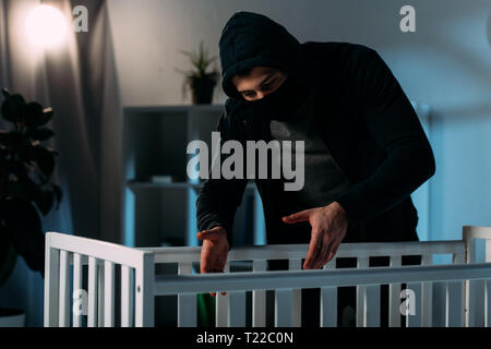 Criminal in mask and black clothes kidnapping child from crib Stock Photo