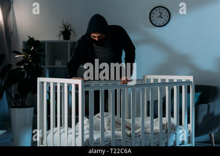 Kidnapper in black clothes standing in dark room and looking in crib Stock Photo