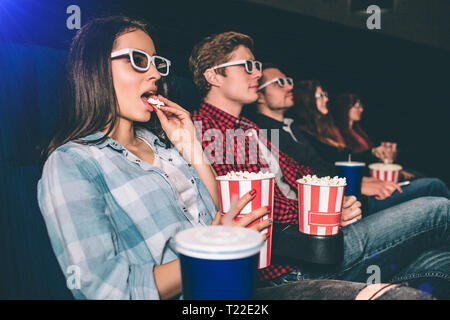 Serious and concentrated people are watching movie. They are sitting in one row. Brunette girl has a basket of popcron and eating it. The other people Stock Photo