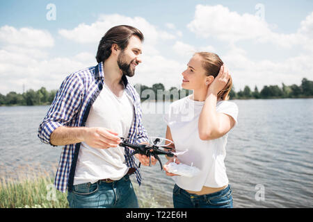 Lovely picture of young man and woman standing together and looking at each other. They are in love with each other. Guy is holding drone in hands. Wo Stock Photo