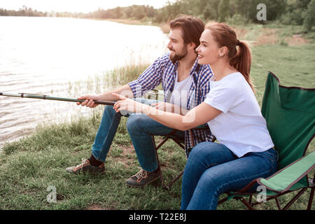 Nice couple is sitting together at the river shore and trying to catch some fish. They are sitting in soft charis and holding fish-rod together. They  Stock Photo