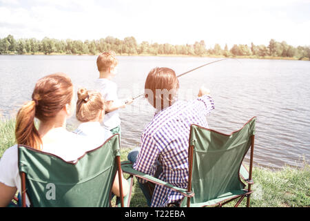 A picture from the back. Small boy is holding a fish-rod and trying to catch some fish in the river. His dad is guiding him and pointing forward. Woma Stock Photo