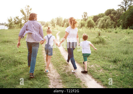 Happy family is walking down the road in meadow. Parents are holding their kids by hands. They are jumping and enjoying the moment Stock Photo