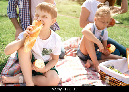 A picture of kids sitting on grass and eating food. Girl is eating an apple while boy is biting bread. Their parents are sitting behind them Stock Photo