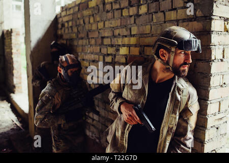 A picture of serious bearded man hiding behind wall and looking to the side. He has gun. Other two men in maska are standing behind him. They are wait Stock Photo