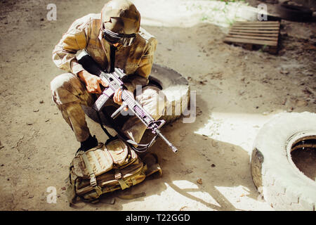 A picture of soldier sitting on the ground and wearing face mask. He is holding black rifle in hands. Man is looking odwn to bag. He has some rest Stock Photo
