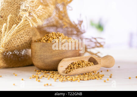 Lot of whole white mustard seeds with wooden bowl , wooden scoop in a jute bag with red flowers in a white kitchen