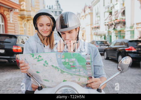 Concentrated and nice people are sitting on motorcycle and looking at map. Guy is holding it. They are standing in middle of road. They wear helmets Stock Photo
