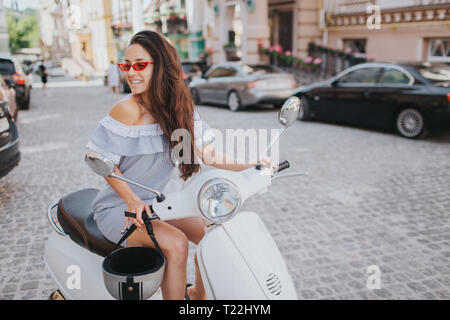 Beautiful and amazing brunette girl is sitting on motorcyle and looking to the right. She wears red glasses. Girl is posing and smiling Stock Photo