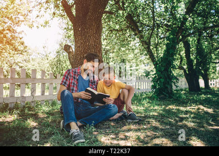 Careful an good father is sitting with his son inder tree and holding book. They are looking at it. They are reading it. Boy is very interested. He lo Stock Photo