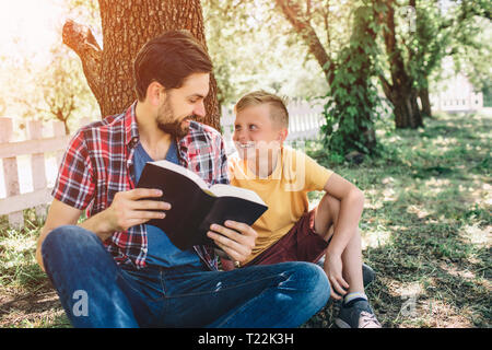 Nice and cheerful father is looking at his son and holding a book. It is opened. Boy is looking at his father and smiling. They are spending time toge Stock Photo