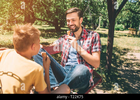 A picture of happy bearded man sitting on blanket with his legs crossed and looking at son. Guy is holding ice cream in hands and smiling. Child is ea Stock Photo