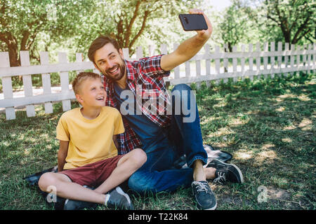 Man is sitting with his so on grass and holding phone in hand. He is taking selfie with kid. They are miling and posing on camera Stock Photo
