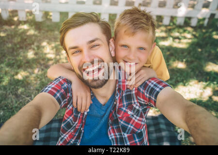 Selfie of happy and bearded guy and his son. Man is holding camera in hands and looking on it. He is smiling. His son is hugging dad and looking on ca Stock Photo