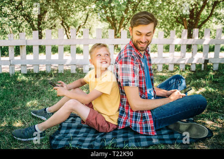 Father is sitting back to back with his son. They are holding phones in hands. Boys are looking at each other and smiling Stock Photo