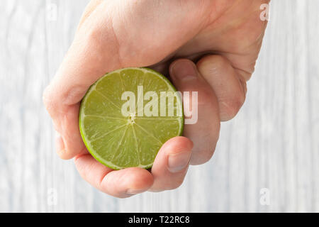 The hand of a man squeezes the juice from a slice of lime half. Juicy drop hanging down. Light wooden background. Stock Photo