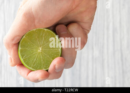 The hand of a man squeezes the juice from a slice of lime half. Juicy drop hanging down. Light wooden background. Stock Photo