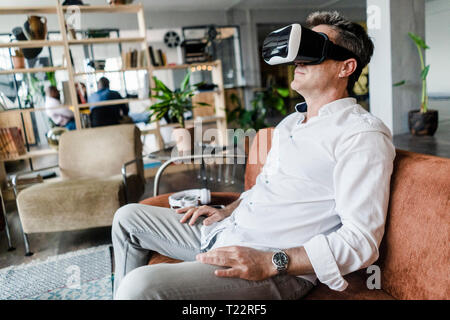 Mature man wearing VR glasses sitting on couch in a loft Stock Photo