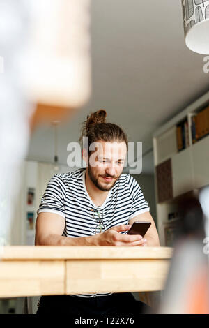 Young man with a bun sitting at home, using smartphone Stock Photo