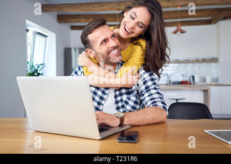 Happy couple sitting at dining table, embracing, using laptop Stock Photo