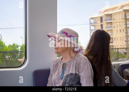 Beautiful gray-haired grandmother in a sunhat rides a commuter train and looks out the window. Stock Photo