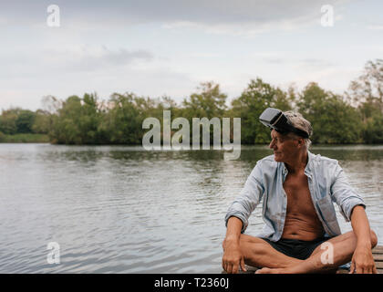 Senior man sitting on jetty at a lake wearing VR glasses Stock Photo