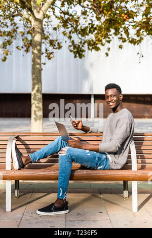 Young man sitting on a bench in the city, using laptop and smartphone Stock Photo
