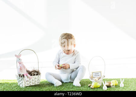 cute kid sitting near straw baskets with Easter eggs, bunnies and happy Easter card isolated on white Stock Photo