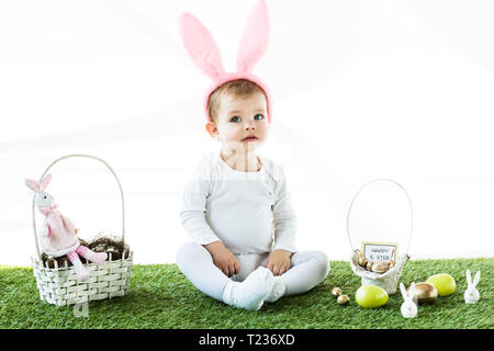 cute baby in bunny ears headband sitting near straw baskets with Easter colorful eggs and decorative rabbits isolated on white Stock Photo