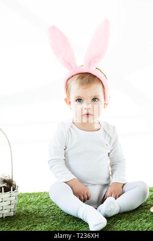 cute dreamy child in bunny ears headband sitting on green grass near straw basket isolated on white Stock Photo