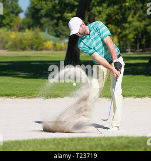 Golfer hits the ball out of sand trap. Focus on golfer, ball and sand wave in motion blur. Stock Photo