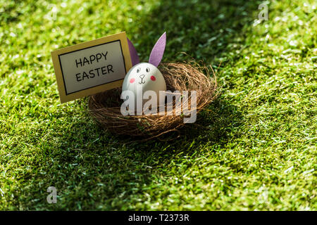 chicken egg with drawn rabbit face and paper ears, and happy Easter card on green grass Stock Photo