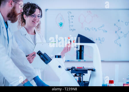Young scientist smiling and standing in front of the microscope in laboratory wearing white coats. Stock Photo