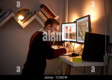 one young man, 20-29 years old, standing in front of a computer while writing in a notebook. night time. Stock Photo