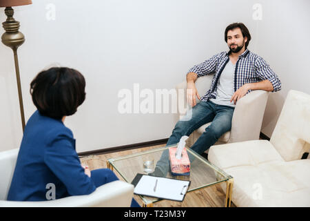 Handsome and relaxed man is sitting in front of psychologist and looking at her. He is very confident. Guy is smiling a bit. He is listening to doctor Stock Photo