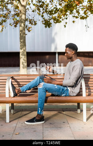 Young man sitting on a bench in the city, using laptop and smartphone Stock Photo