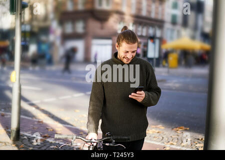 Smiling young man with bicycle in the city looking at cell phone Stock Photo