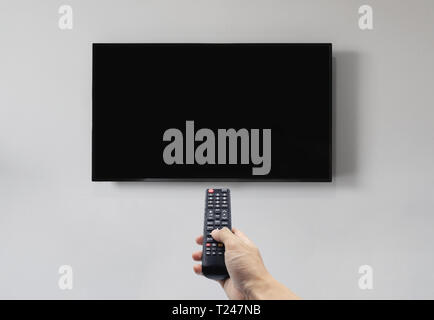 Television on white wall with hand using remote control, blank black screen Stock Photo