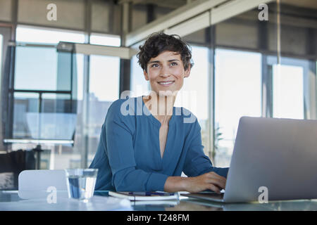 Smiling businesswoman sitting at desk in office with laptop Stock Photo